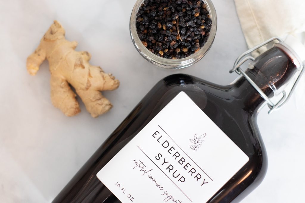 elderberry syrup bottle laying on marble countertop with ginger root and dried elderberries