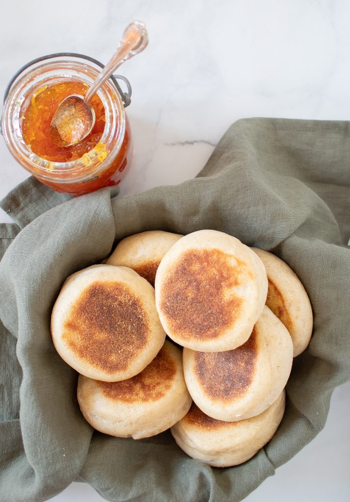 linen basket fully of golden fluffy sourdough English muffins with a jar of orange marmalade sitting beside