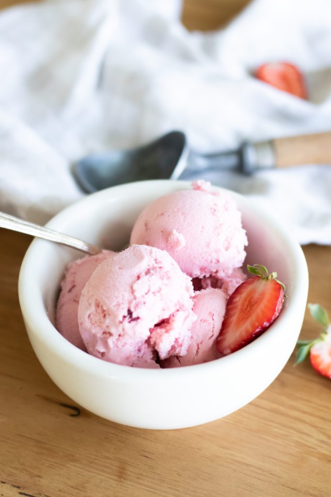light pink raw milk strawberry ice cream in a while bowl with spoon sitting on kitchen table garnished with strawberry
