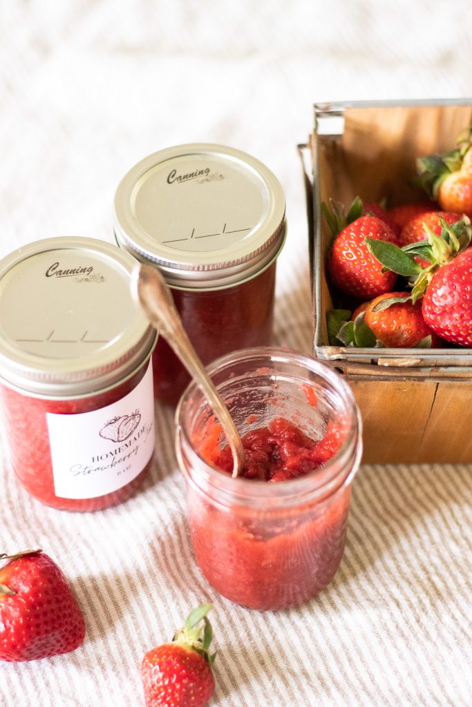 Fresh strawberry jam in jar with spoon next to canned strawberry jam and a quart of fresh strawberries