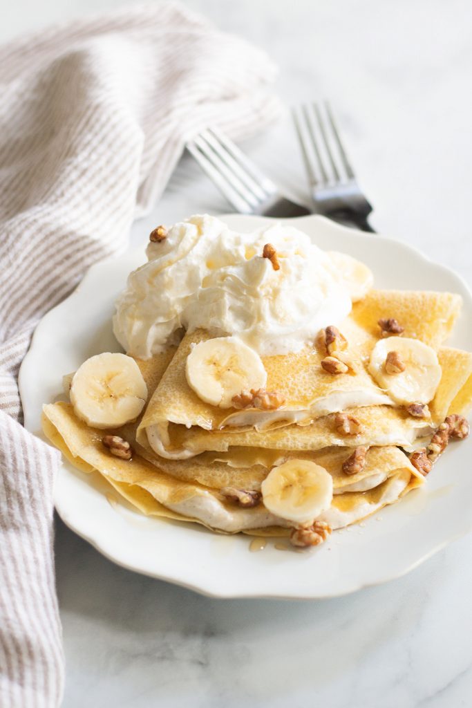 Sourdough Discard Crepe served on white plate with bananas, walnuts, whip cream and syrup.